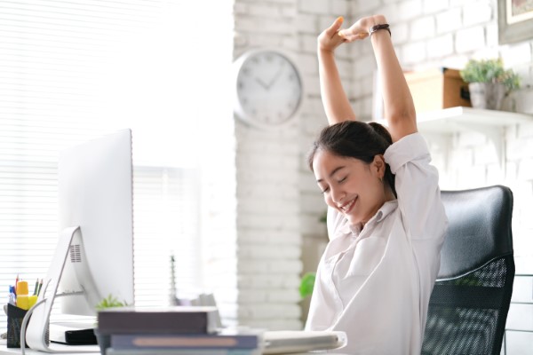 A woman stretching and smiling at her computer desk.
